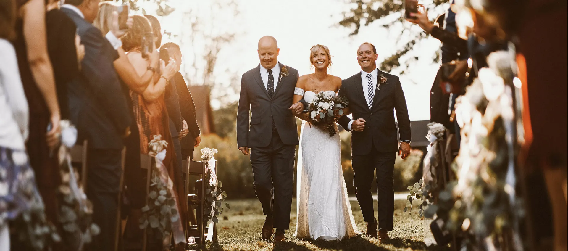 Bride walking down an outdoor aisle between two men as guests film her in golden light at Bishop Farmstead, New Jersey