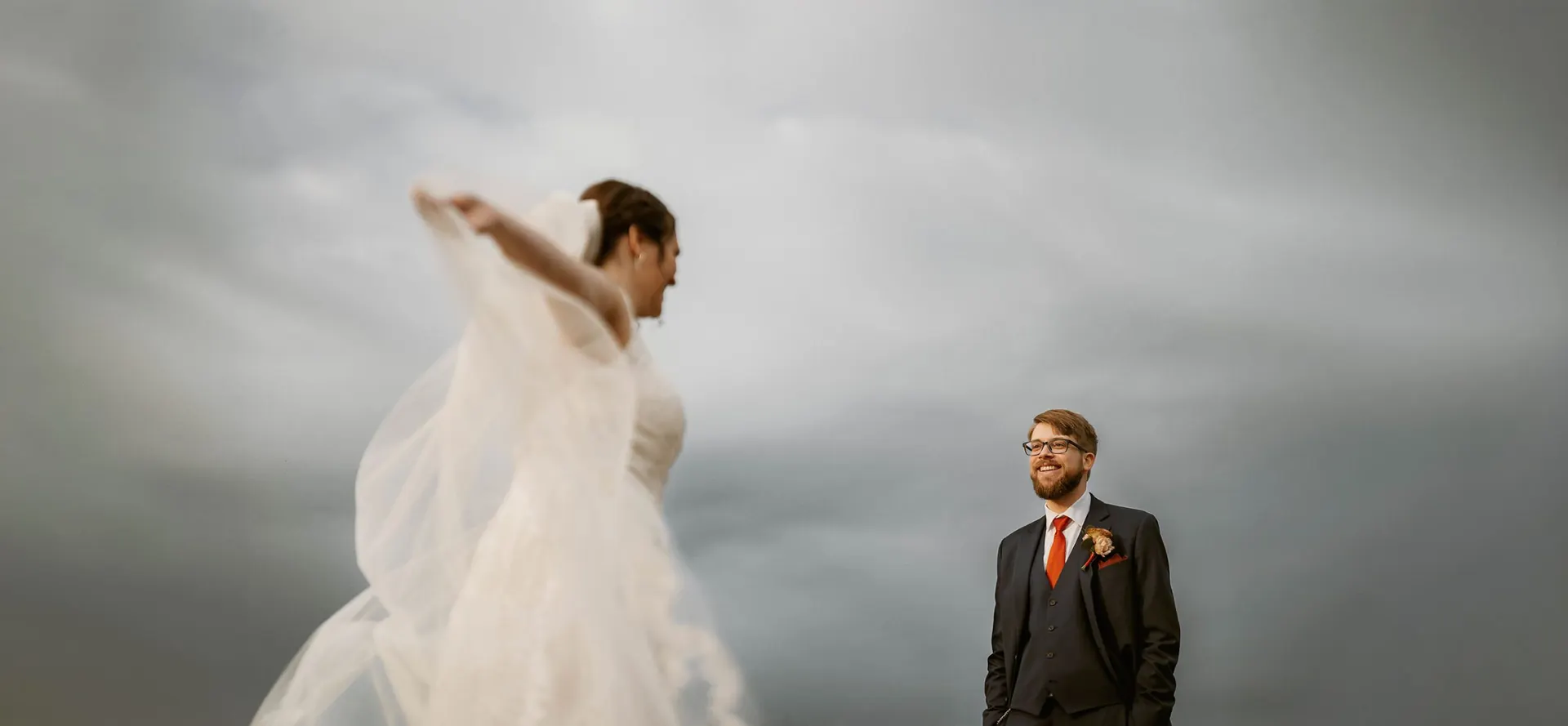 Bride tossing her veil in the wind while the groom smiles under a stormy sky at Bishop Farmstead, New Jersey