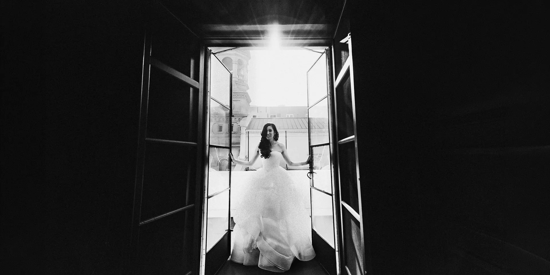Black and white photo of a bride in a full gown opening a pair of balcony doors in Los Angeles