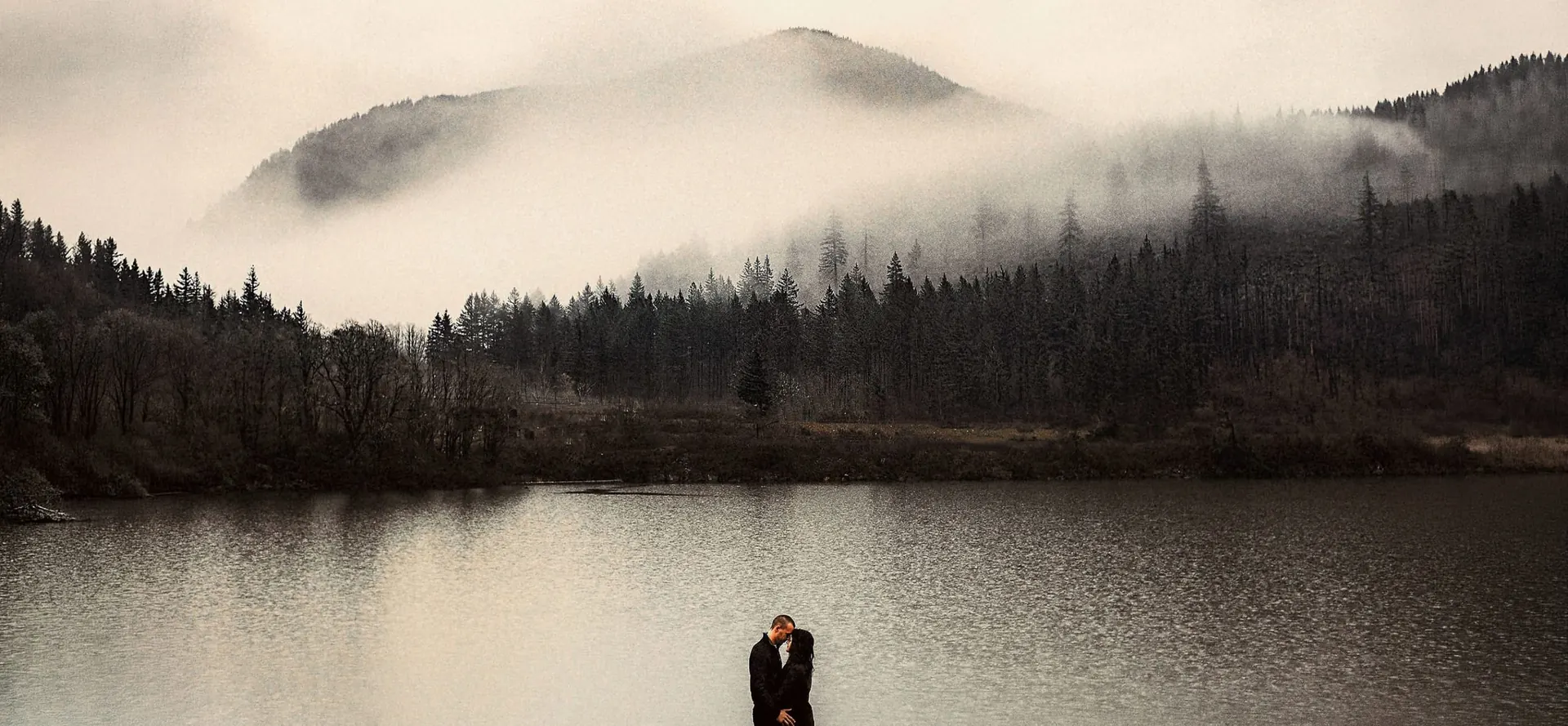 Couple sharing a quiet moment by a lake with fog-covered mountains behind them in Oregon