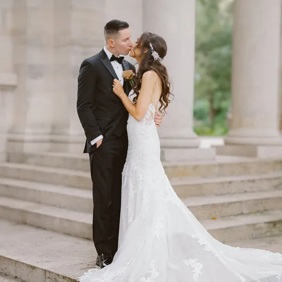 Bride and groom kissing on stone steps between classical columns, her lace train spilling down the stairs