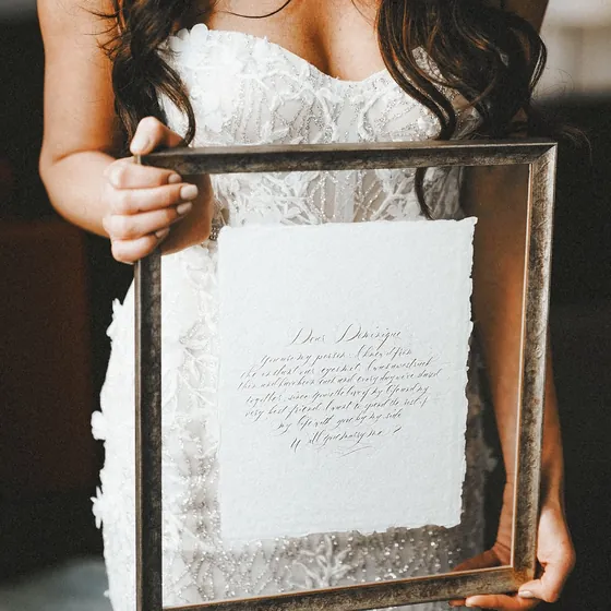 Bride in a beaded lace gown holding a framed calligraphy proposal letter