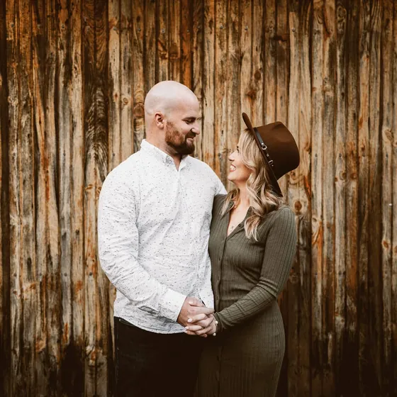Engaged couple holding hands and smiling at each other in front of a weathered barn wood wall