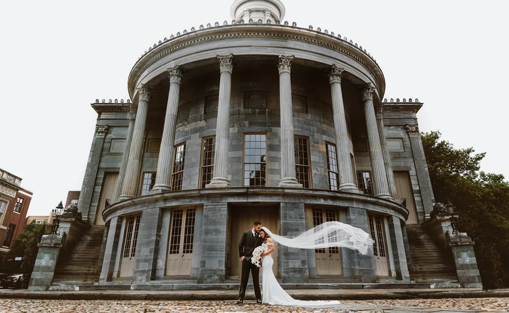 Bride and groom embracing as her veil flows in the wind outside the Merchants' Exchange Building in Old City Philadelphia