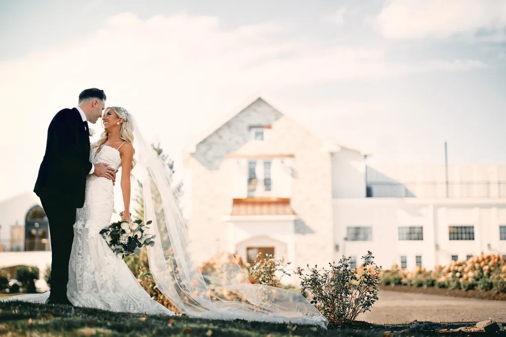 Smiling bride and groom with her long cathedral veil trailing across the lawn at Renault Winery in Egg Harbor City, New Jersey