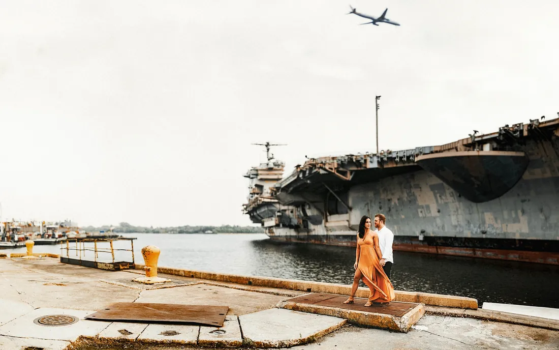 Engaged couple on a pier beside an aircraft carrier as a plane flies over