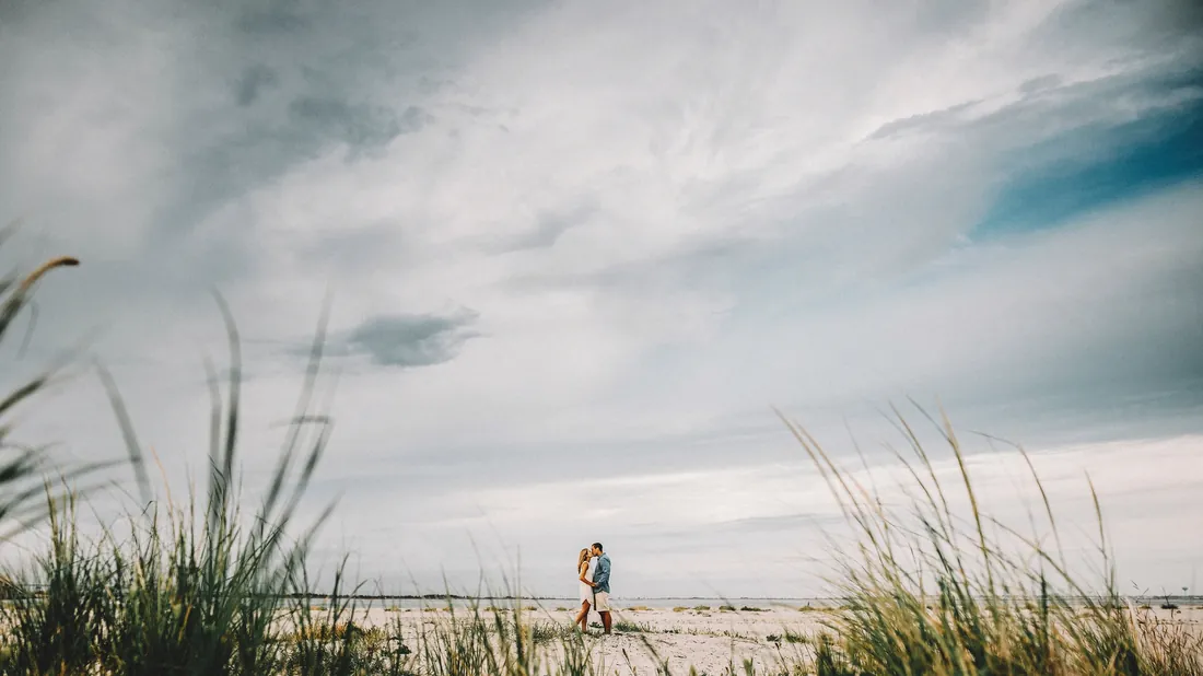 Engaged couple small on the beach beyond the dune grass