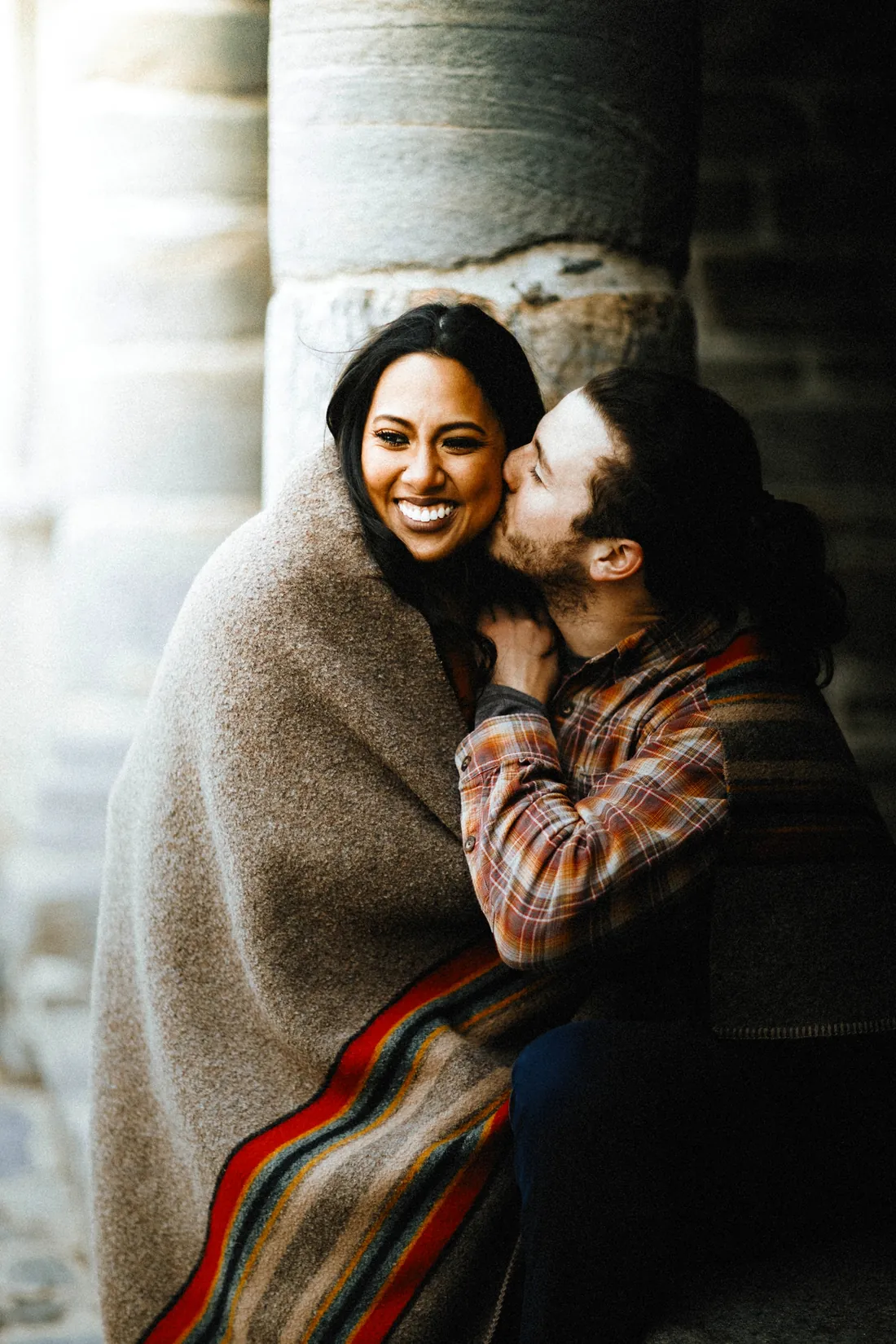 Engaged couple wrapped in a blanket, laughing and kissing