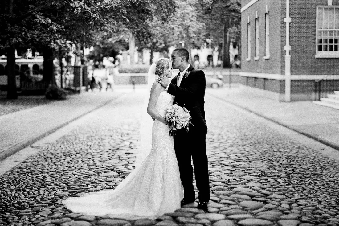 Black and white film photograph of a bride and groom kissing on a cobblestone street