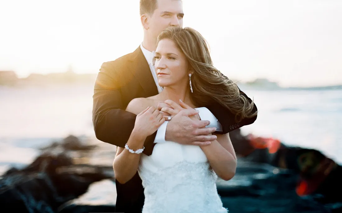 Film photograph of a groom holding his bride from behind by the sea at sunset