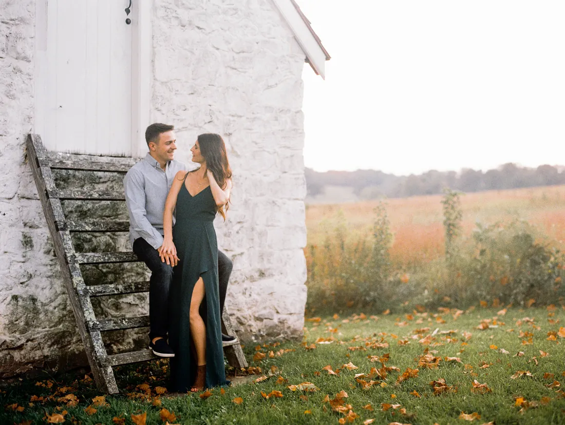 Film photograph of an engaged couple leaning against a white stone house beside a field