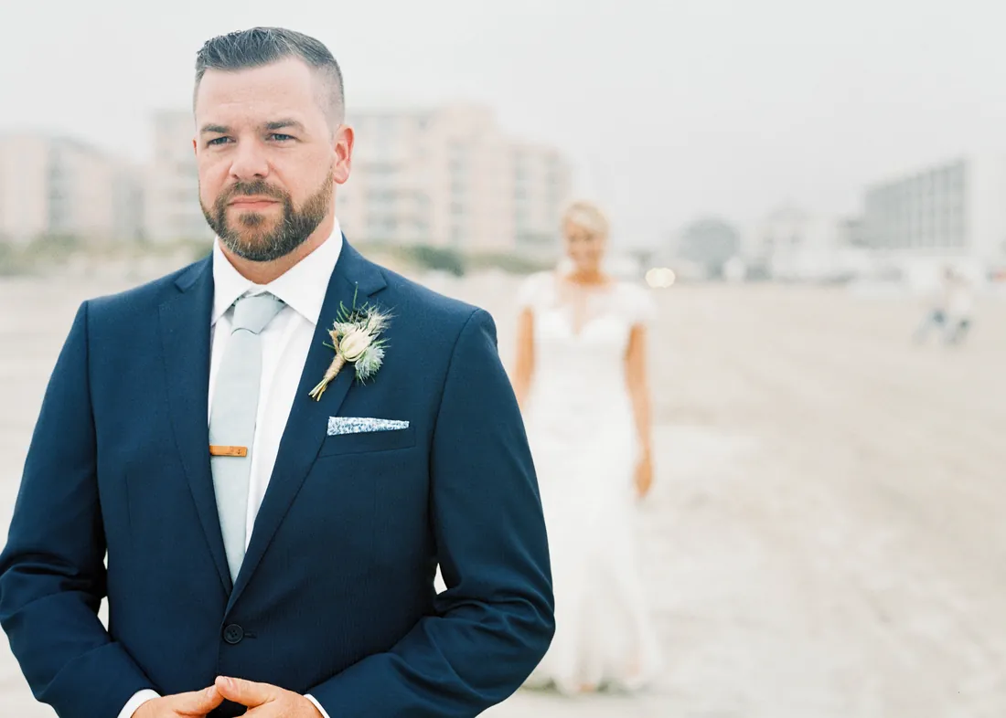 Film portrait of a groom on the beach with his bride walking up behind him
