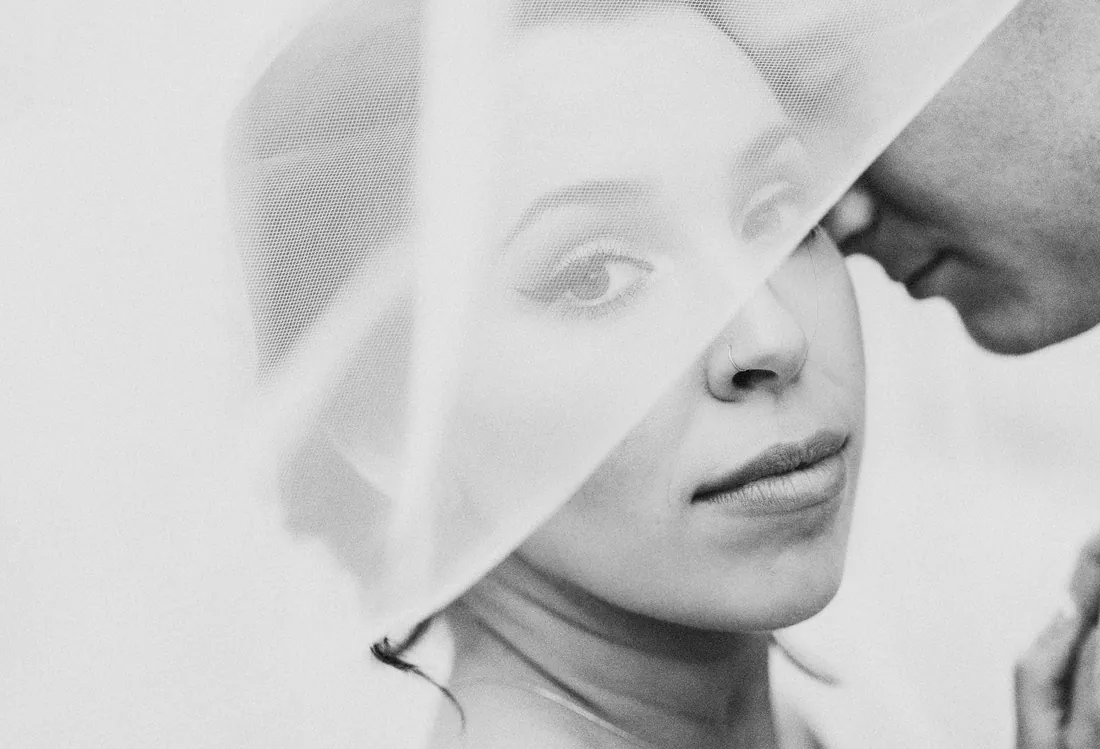 Black and white portrait of a bride looking through her veil as her groom leans in