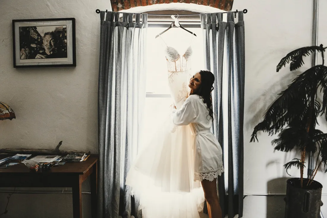 Bride in her robe with her dress hanging in the window