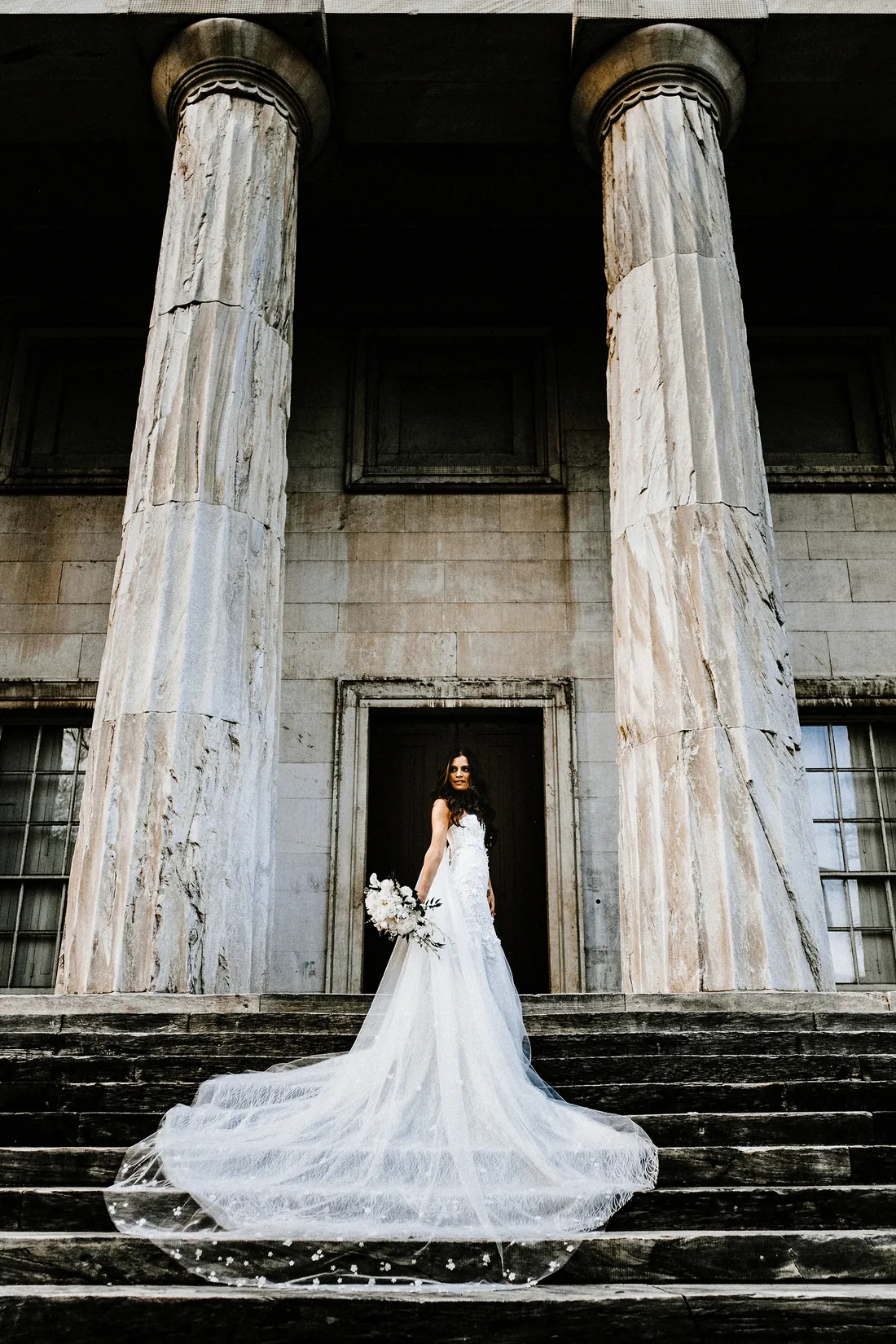 Bride between tall stone columns with her train spread down the steps