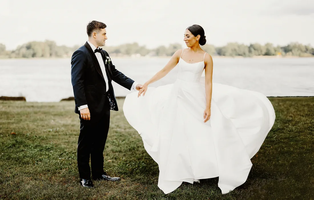 Bride swishing her gown as her groom watches by the water