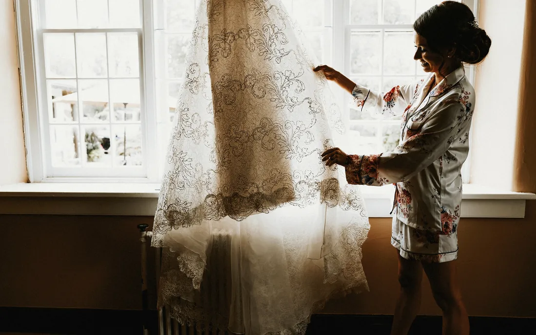 Bride in her robe reaching for her lace gown by the window