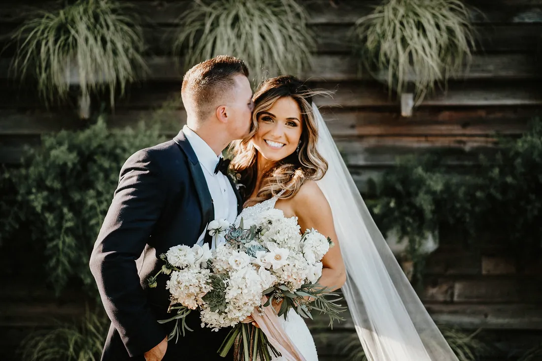 Groom kissing his laughing bride on the cheek in front of a wall of hanging plants