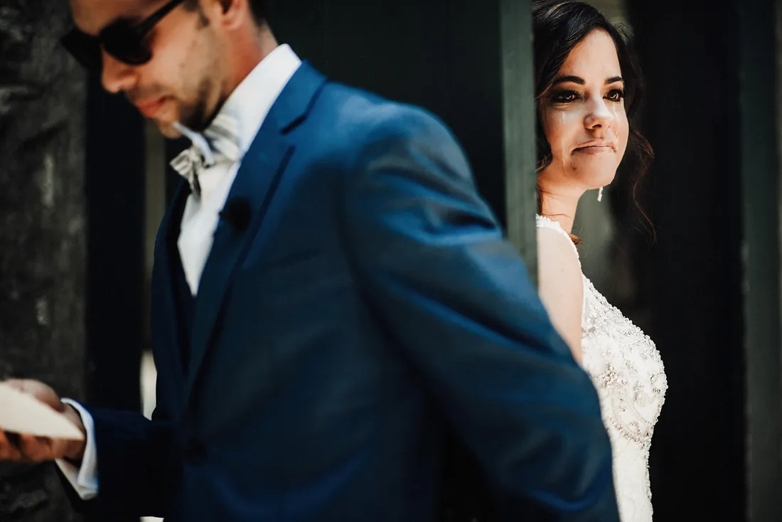 Groom in sunglasses reading a letter as his bride waits behind the door