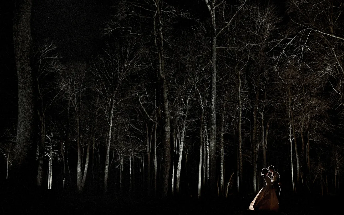 Bride and groom small at the edge of a dark forest at night