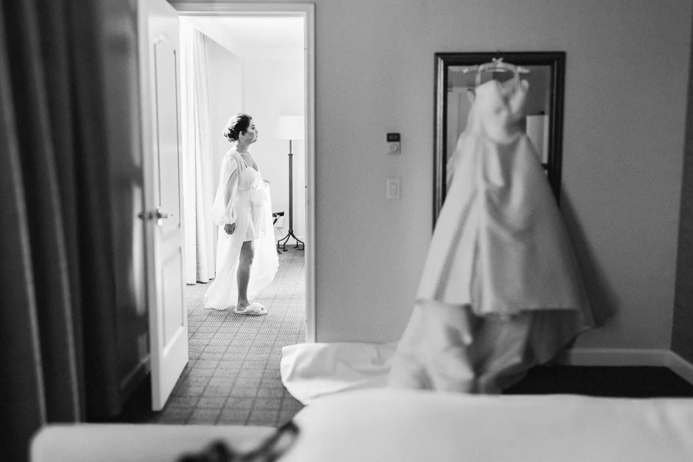 Black and white photo of Celeste in her robe looking at her wedding dress hanging in the room