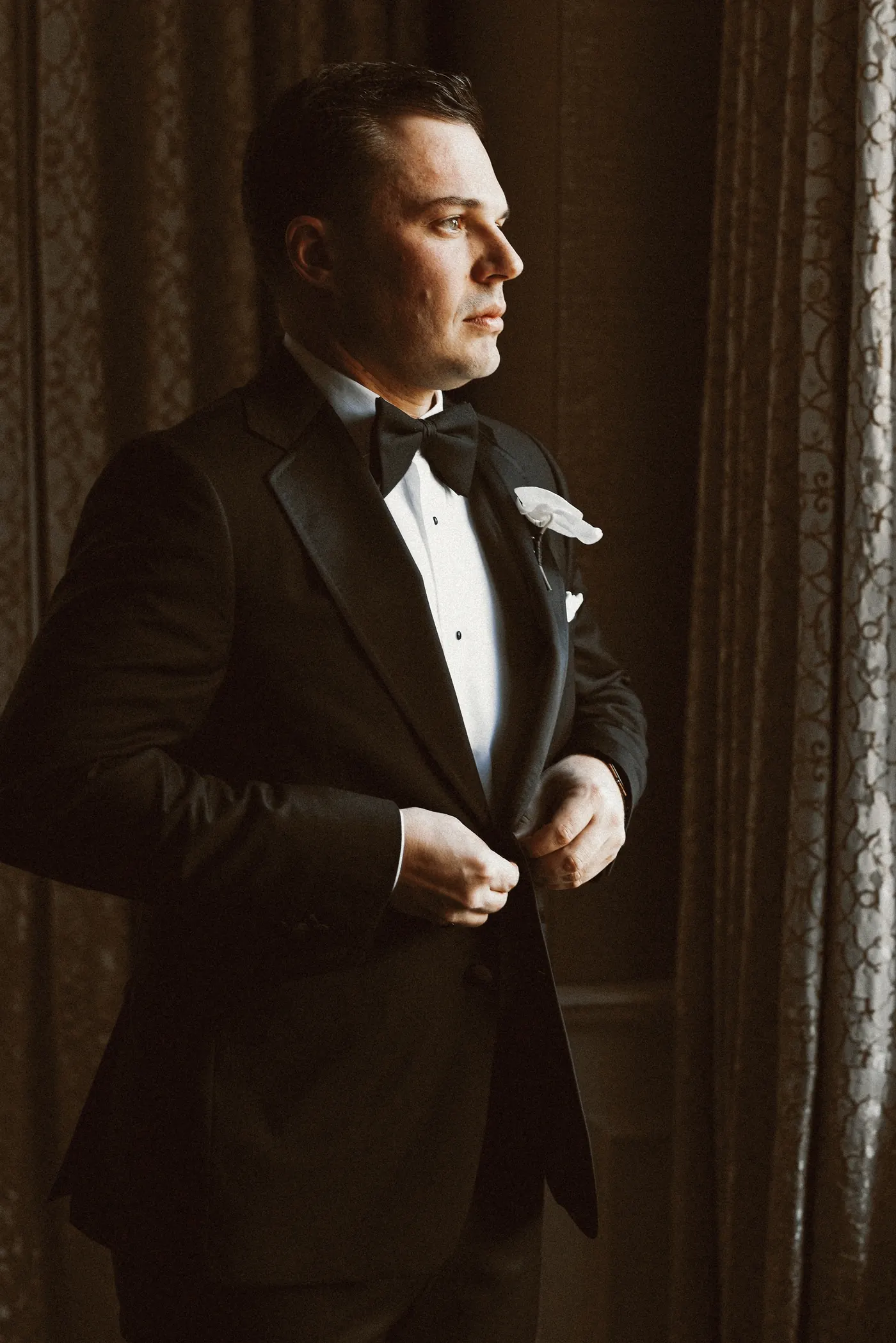 Branon buttoning his tuxedo jacket by the window
