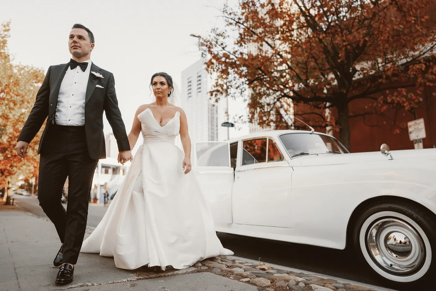 Branon and Celeste walking beside the white vintage car under fall leaves in Center City Philadelphia