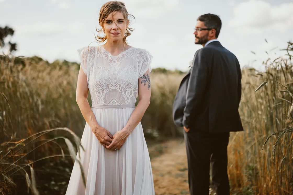 Erica in a lace wedding dress in a tall grain field with Matt behind her at Johnson's Locust Hall Farm