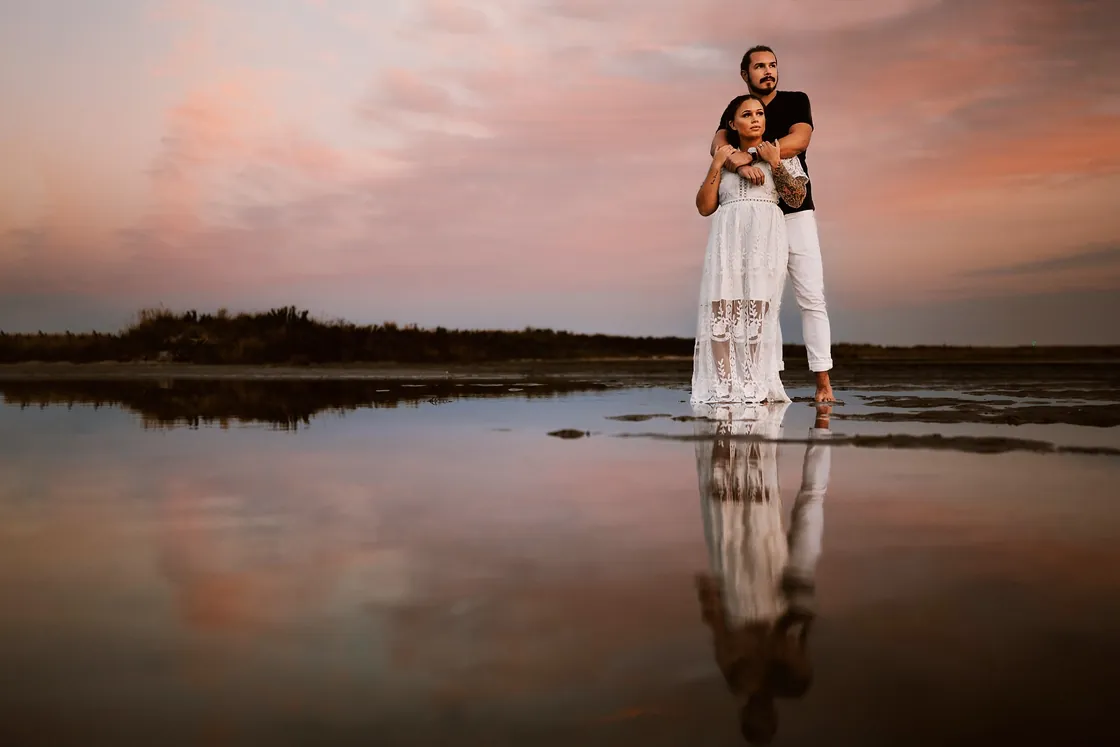Dan and Madison embracing at the water's edge under a pink sunset during their engagement session at Barnegat Lighthouse on Long Beach Island