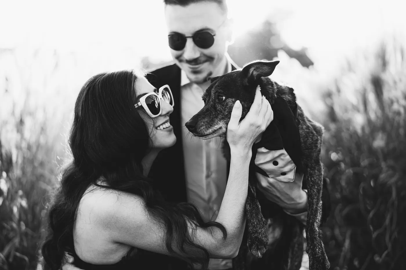Black and white photo of Morgan laughing as Will holds their dog in a tuxedo bib